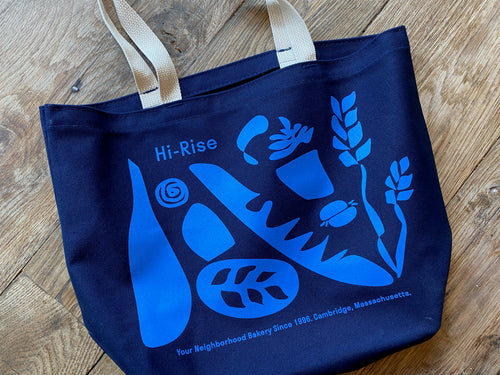 Dark Blue Tote with light beige handles. Tote bag has loose abstract designs of a wine bottle, cheddar snail, wheat, sandwich, baguette, loaf of bread, and a plant. Top left text reads 